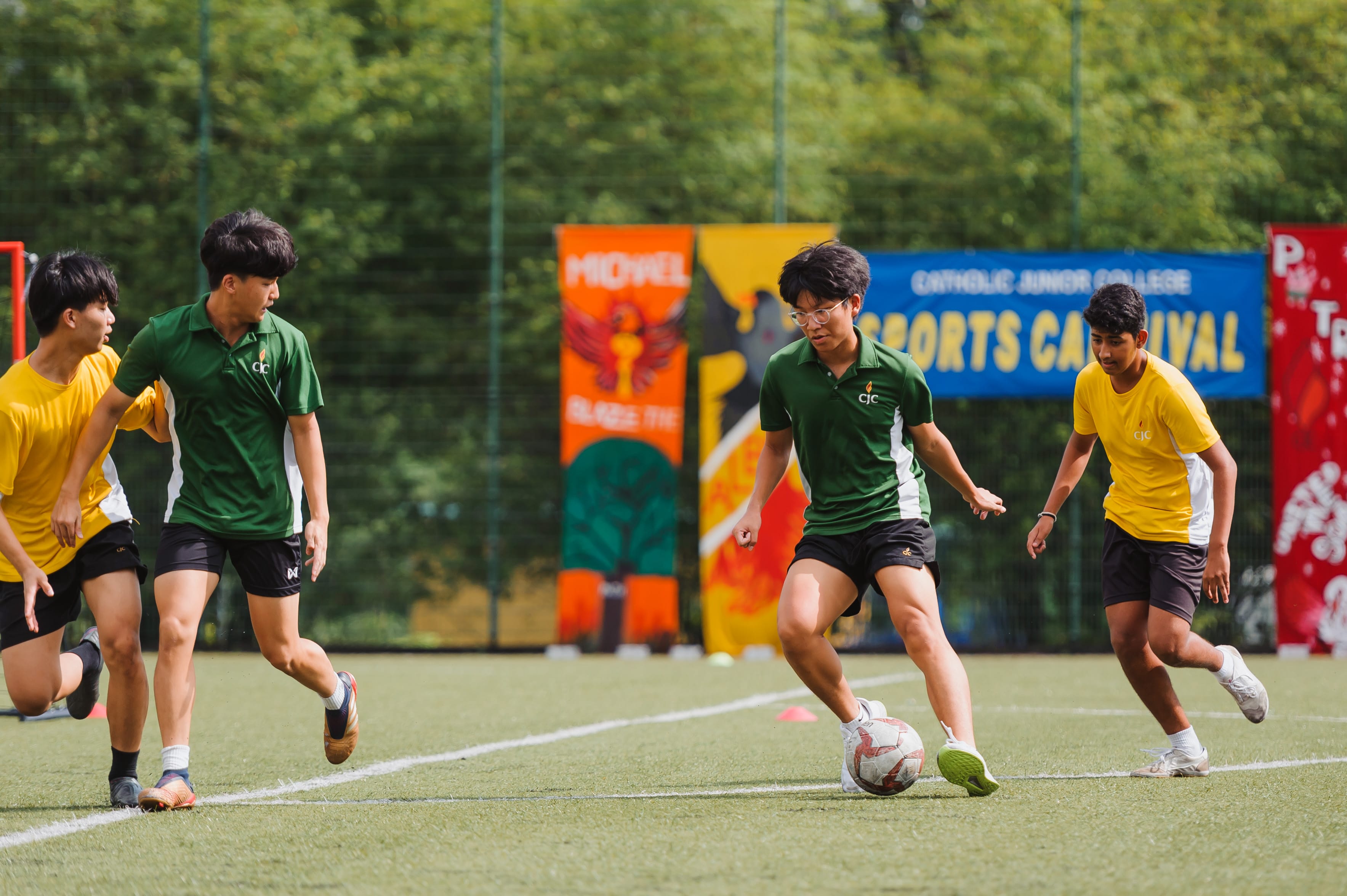 Four teenage boys play football on a green field at a Catholic Junior College Sports Carnival event.